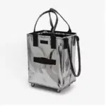 Uma grande bolsa de compras com zíper com zíper em prata brilhante, Apresentando alças e rodas pretas, fica de pé em um fundo branco.