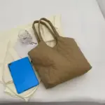 Uma bolsa acolchoada inchada para mulheres em marrom, um comprimido azul, e um vidro transparente é colocado em um fundo de pano creme.