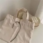 Borża tax-xiri tal-kanvas li tiġbed fil-beige bi spag iswed, customizable permezz ta 'OEM u ODM, titqiegħed fuq mejda bajda ħdejn platt li żżomm xemgħa tal-bżieżaq.