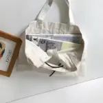 Borża tax-xiri tal-kanvas li tiġbed bil-kulur tal-krema mimlija rivisti tistrieħ fuq wiċċ abjad ħdejn stampa inkwadrata.