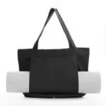 Women's Yoga Fitness Tote with shoulder straps, zobrazené čiernou farbou, Drží v prednom vrecku zvinutú rohož na jogu na bielom pozadí.