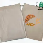 രണ്ട് ബീജ് കോട്ടൺ ബ്രെഡ് ഡ്രോസ്ട്രിംഗ് ഷോപ്പിംഗ് ബാഗുകൾ; ഒരാൾക്ക് ഒരു ക്രോസന്റ് ഉണ്ട്, ഗോതന്വ്, and "LOGO" അച്ചടിക്കല്, ഓം / ഒഡിഎം ഓപ്ഷനുകൾ ഹൈലൈറ്റ് ചെയ്യുന്നു. മുകളിലെ വലതുവശത്ത് ഹോണൻസ്ക് ലോഗോ ദൃശ്യമാകുന്നു.